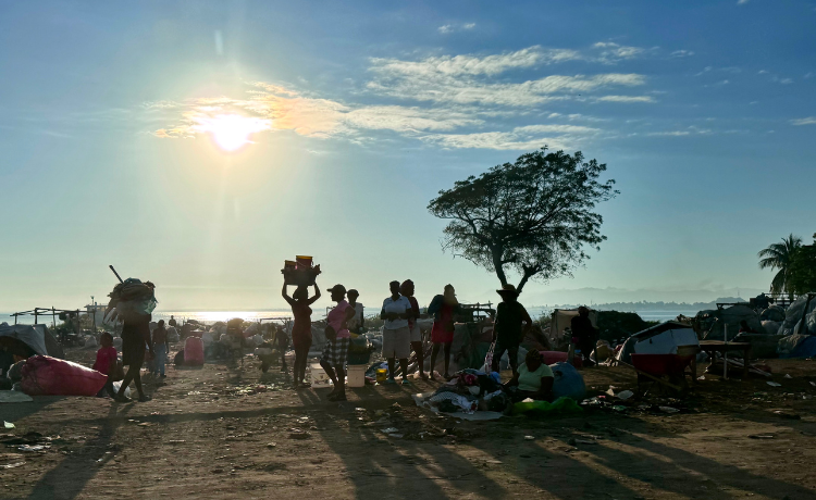 As the sun sets over the ocean, a group of people stand backlit by the side of a dirt road surrounded by their belongings