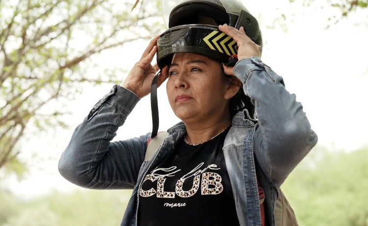 A woman in a denim jacket holds a motorcycle helmet over her head.