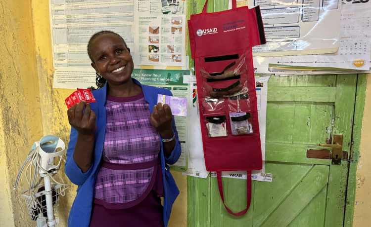 A woman in a blue blazer and purple dress smiles as she holds up male and female condoms in a clinic. Next to her is a USAID-logoed display of contraceptive methods.