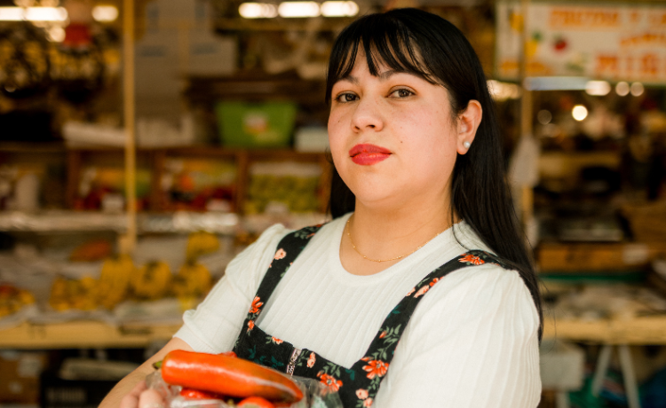 A woman with dark hair and wearing a flowery top over a white t-shirt holds a bag of red peppers in front of a market stall