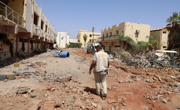 A man in a UNFPA vest walk among the rubble of a street with destroyed buildings along it