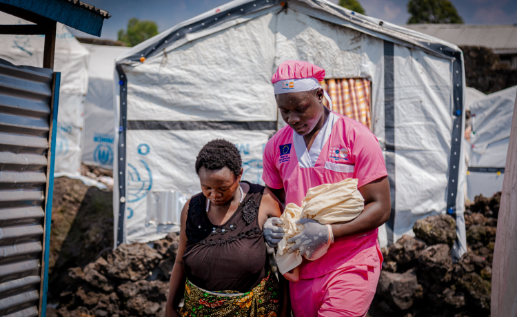A male health worker in pink scrubs accompanies a pregnant woman in a displacement site with white tents visible in the background