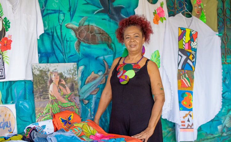 A woman in a black dress and bright jewellery stands before a selection of patterned clothes and paintings