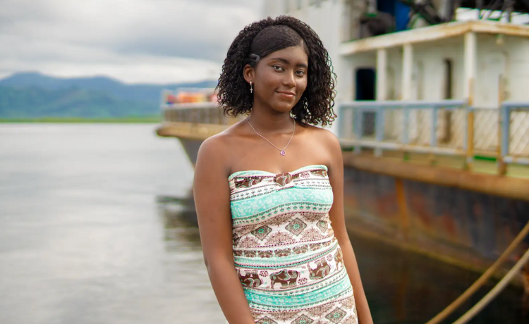 A young woman in a turquoise and brown pattered dress stands smiling beside a boat