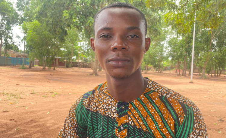 A man in a green and orange patterned shirt smiles at the camera. He is in a dusty field with trees in the background