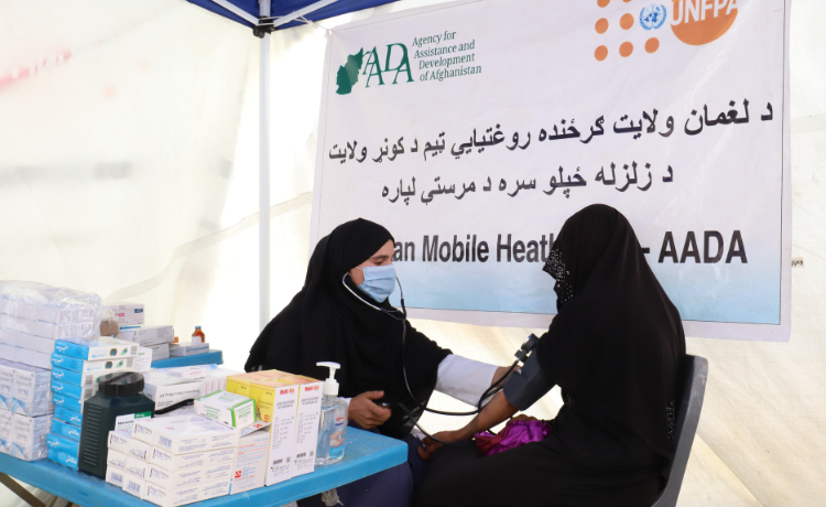 A female health worker takes the blood pressure of a young woman while seated in front of boxes of medicine inside a white UNFPA tent