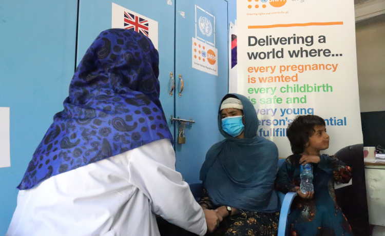 A female health worker assists a woman seated beside a child, with a UNFPA banner visible behind her