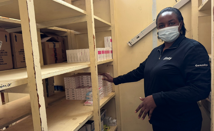 A pharmacy stands between wooden shelves, touching boxes on one of the shelves. Several shelves above her and beside her are empty.
