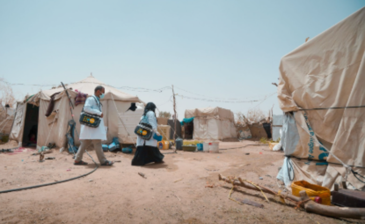 Two health workers with UNFPA-branded medical bags walk through a displacement camp