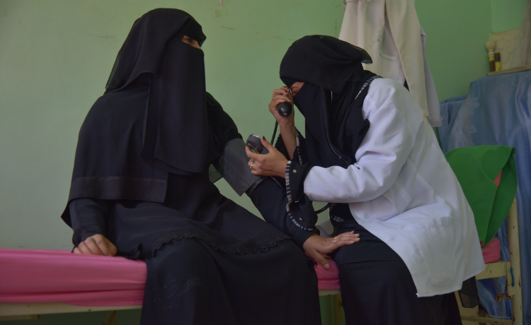 A female health worker sits on a hospital bed assessing a female patient in a ward