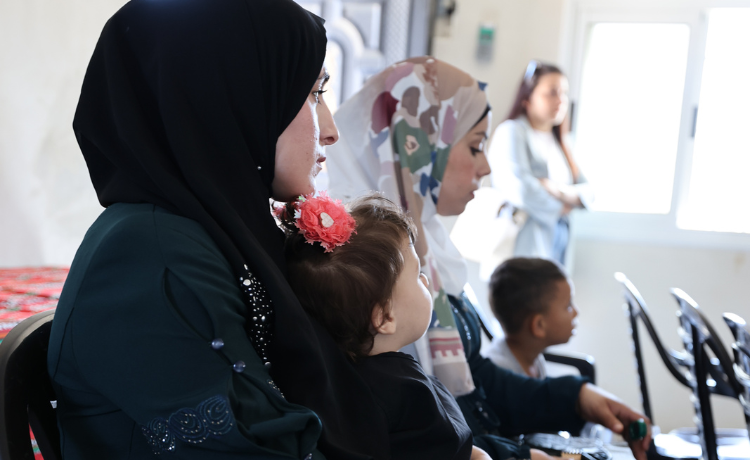 A woman sits on a chair inside a pale room, holding a young girl on her lap