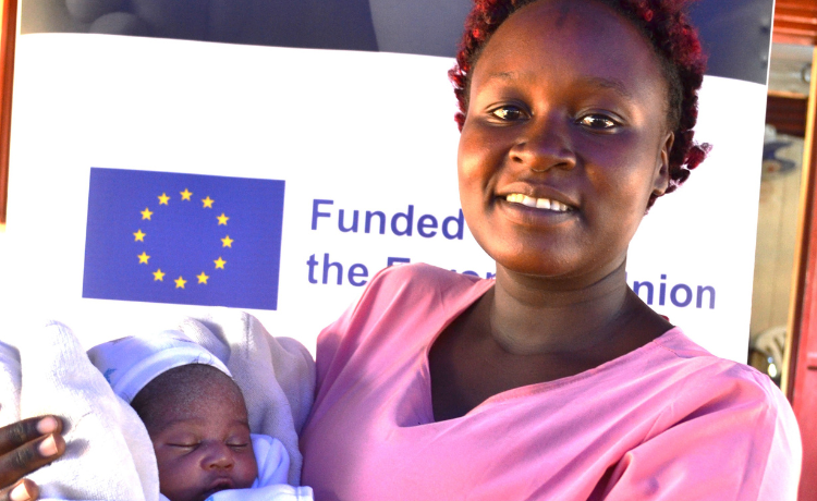 A woman in pink holds her newborn baby. She is standing in front of a poster for Safe Birth Africa.