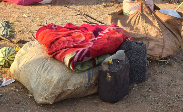 A blanket, two jerrycans, two canvas bags and a melon are placed on the sandy ground outside a tent.