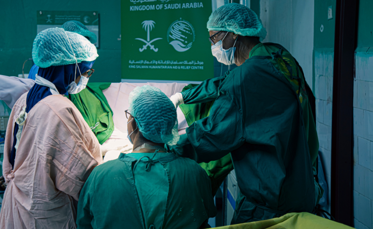 Three health workers in surgical garb are seen from the back as they perform a surgery. The person receiving the surgery is not visible.