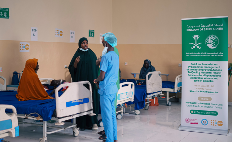 A recovery room is lined with hospital beds and blue linens. A woman with a face mask speaks to two health workers in scrubs. A banner shows the logo of KSRelief.