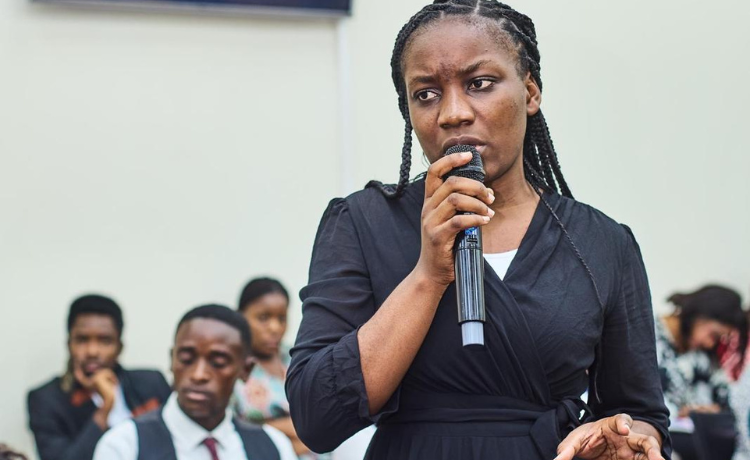  A young woman speaks into a microphone in front of a seated crowd