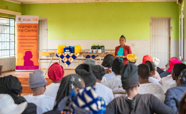 A woman in a red blazer stands before a crowd of women inside a brightly lit green-painted room.