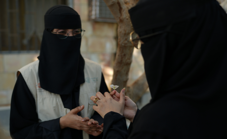  A woman in a UNFPA vest, black niqab and glasses speaks with another woman dressed in black, who is seen only from behind