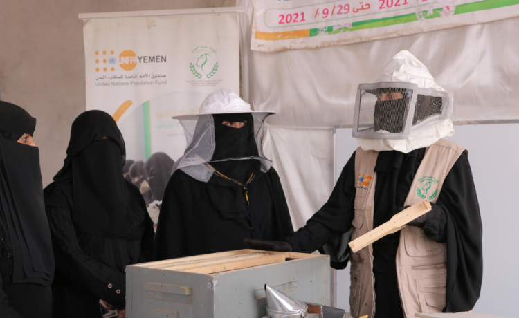 Women in protective beekeeping masks speak to other women inside a white room