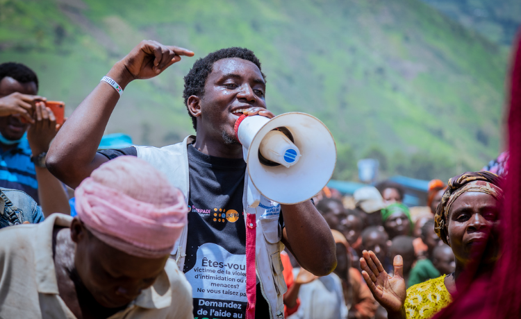  A man in a UNFPA t-shirt holds a loudspeaker and addresses a crowd of people; green hills can be seen in the background
