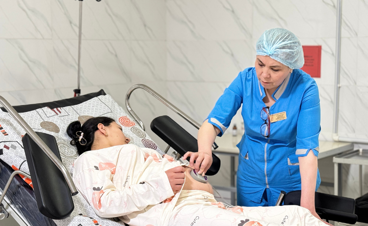 A health worker in blue scrubs and surgical cap places an electronic fetal monitor on a patient wearing pajamas, resting in an adjustable hospital bed. They are in a brightly lit room with white tiled walls.
