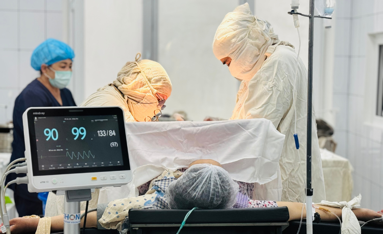 A surgical procedure in a bright operating room, showing a patient on the table, a vital signs monitor displaying '99' and a blood pressure of '133/84', and two surgeons in full protective gowns and head wraps working over the patient. A third healthcare worker stands in the background