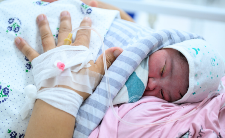 Close-up of a newborn baby sleeping on its mother's chest. The baby wears a white hat and is wrapped in a striped blanket. The mother's hand, which has an IV cannula secured on the back, rests near the baby