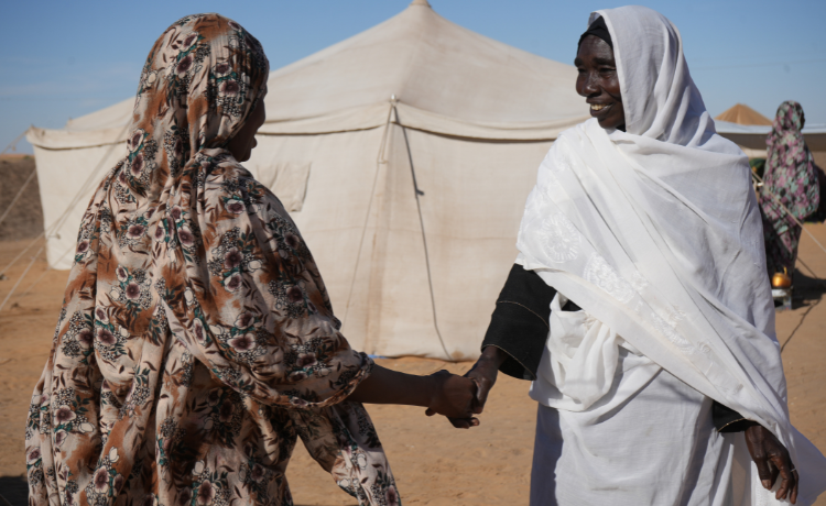 Standing on dusty ground in front of a white tent, a woman dressed in white smiles and shakes the hand of another woman seen from the back