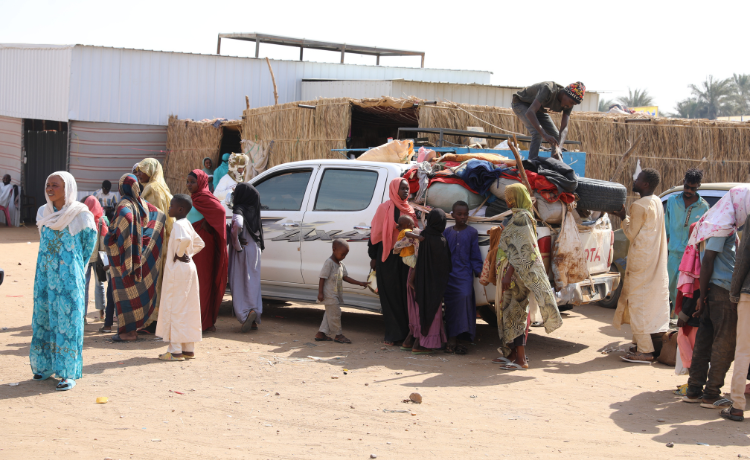 A group of people stand on a dusty road around a white pick-up truck that is crammed full of suitcases and other belongings