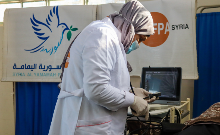 A health worker in a white coat with UNFPA logo visible on the back, wearing a beige headscarf and a blue face mask, examines an ultrasound machine