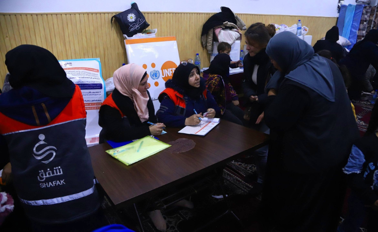  Two women seated behind a dark wooden desk fill in forms and take details from displaced women and their families