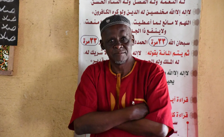 An older man stands inside with his arms crossed, wearing a deep red caftan and a gray patterned cap, looking toward the camera with a calm expression.
