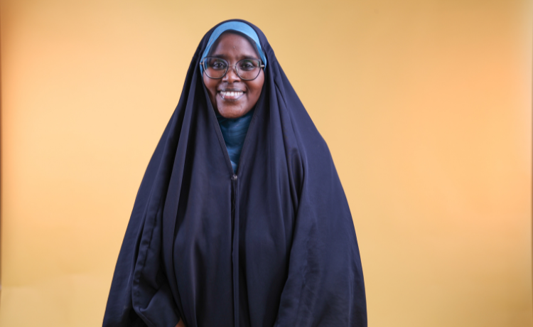  A woman wearing a large black abaya and a light blue hijab stands against an orange background, smiling. She is also wearing glasses, and her hands are clasped in front of her.