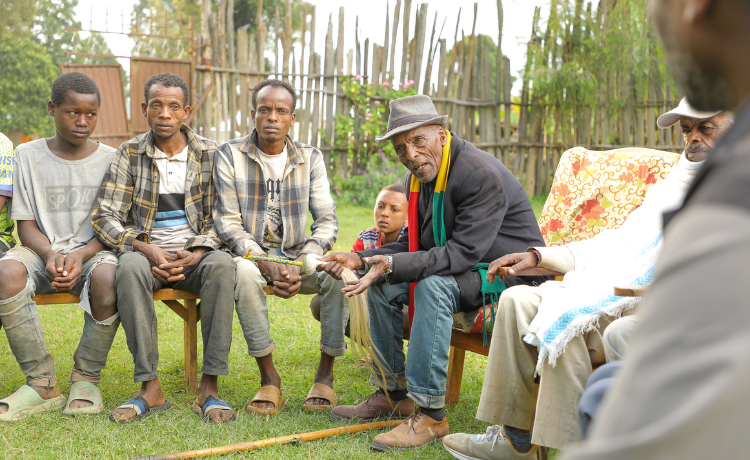  A group of men and one child are seated outside on wooden benches and chairs in a grassy area, engaged in a discussion. An older man wearing a dark jacket, jeans, and a fedora with a black, yellow, and green scarf around his neck is speaking and gesturing with his hands.