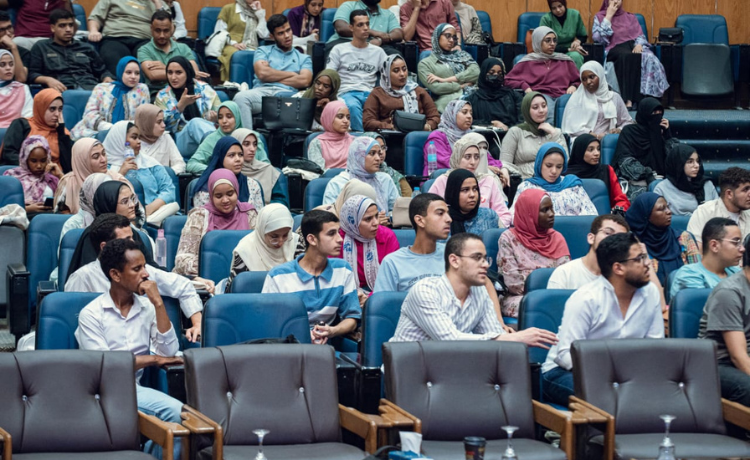 A large group of young adults, both male and female, are seated in an auditorium or lecture hall. Many of the women are wearing hijabs in various colours. They are sitting in rows of blue seats with brown wooden arm rests, and most of the attendees are looking toward the front of the room, suggesting they are listening to a speaker or presentation