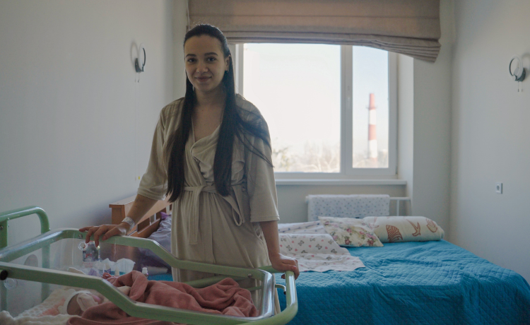 A young woman stands smiling in a bright hospital room, looking towards the camera. She is wearing a light-colored robe and is leaning on the rail of a clear, wheeled crib where a newborn baby is visible, mostly covered by a pink blanket. Behind her is a bed with blue covers, and through a large window, a red and white striped industrial chimney is visible in the distance.