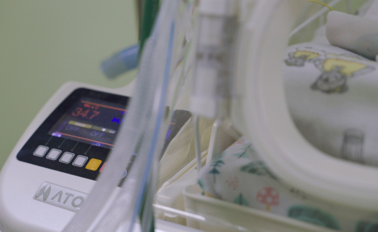 A close-up image of a medical device shows a digital screen displaying "34.7" and the word "OFF" in multiple locations. The control panel is branded with the "ATOM" logo. To the right of the panel, a baby is visible inside an incubator, wrapped in a blanket with a light-coloured, patterned print