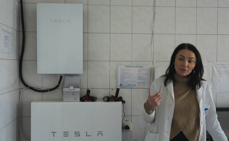 A woman in a white lab coat stands in a room with white tiled walls, gesturing with her right hand. To her left, there are two large white units mounted on the wall, both bearing the TESLA logo. There is text on the wall in what appears to be a Cyrillic script