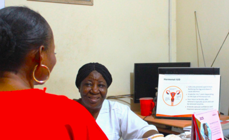 A woman facing away from the camera dressed in a red top consults a healthcare provider wearing a white top. On the desk there are visible information materials and a calendar