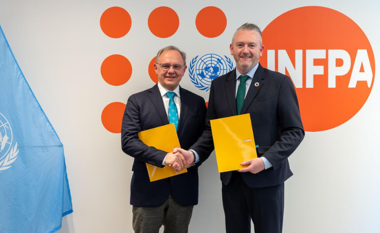 Prof. Frank Louwen and Pio Smith seated at a table signing a document during a formal agreement ceremony, with officials present in the background.