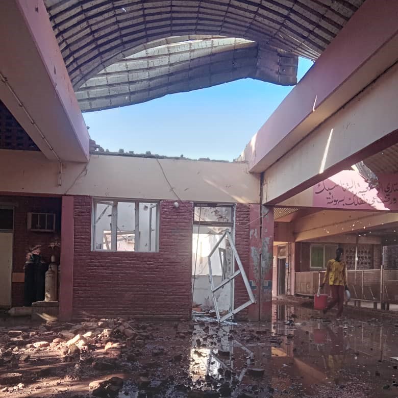  Una persona con camiseta amarilla carga un cubo rojo entre escombros y charcos. El edificio carece de techo