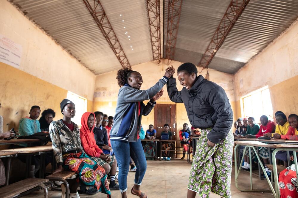 Inside a concrete-walled building with a corrugated iron roof, two young women laugh and hold hands in the air, surrounded by seated young people who are watching them]