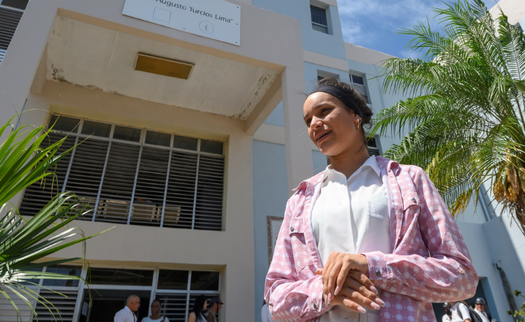 Una joven con camisa blanca y chaqueta rosada frente a una clínica de salud en Cuba.]