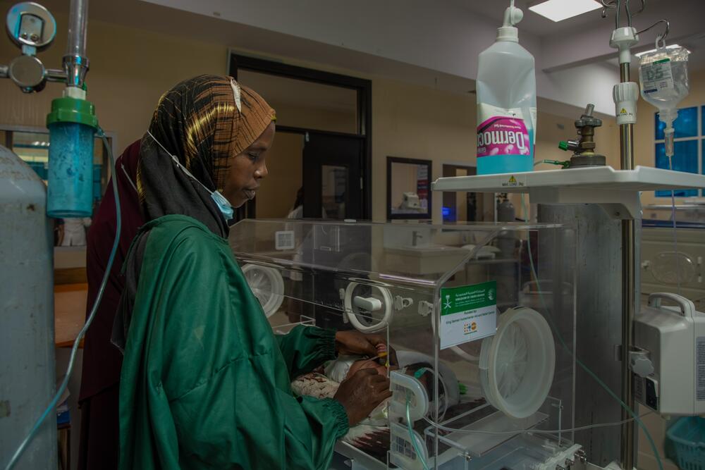 A woman in a brown head scarf and green scrubs looks into an incubator inside a hospital ward