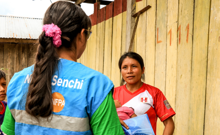 Deux femmes à l’extérieur d’une maison en bois. L’une d’entre elles porte un gilet bleu de l’UNFPA, l’autre tient un jeune enfant dans ses bras