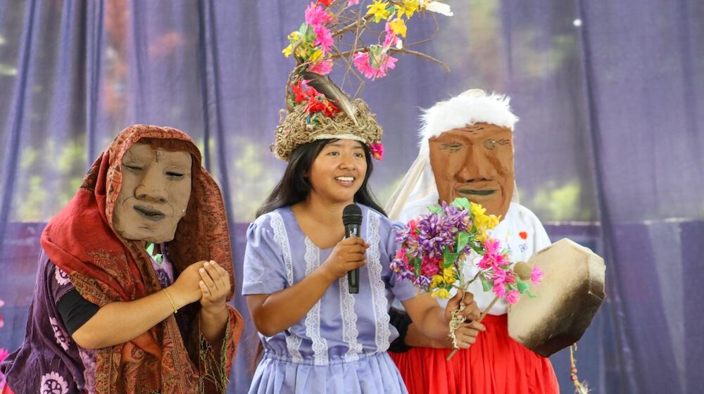 Three women participate in a theatrical performance. The woman in the centre, wearing a lavender dress and a floral headpiece, holds a microphone and a bouquet of colourful flowers. On either side of her are two women wearing traditional clothing and large handmade masks. The woman on the left wears a purple outfit with a patterned red shawl, while the one on the right wears a white blouse and holds a drum. They stand against a purple-toned backdrop.