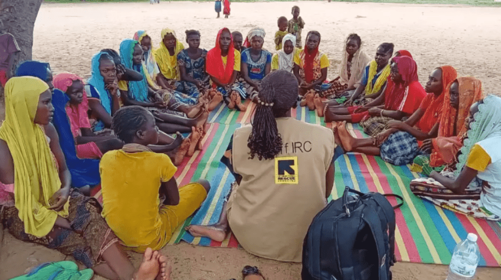 A woman in a vest with the International Rescue Committee logo guides a discussion on a colorful blanket. Women and girls sit in a circle atop the blanket.