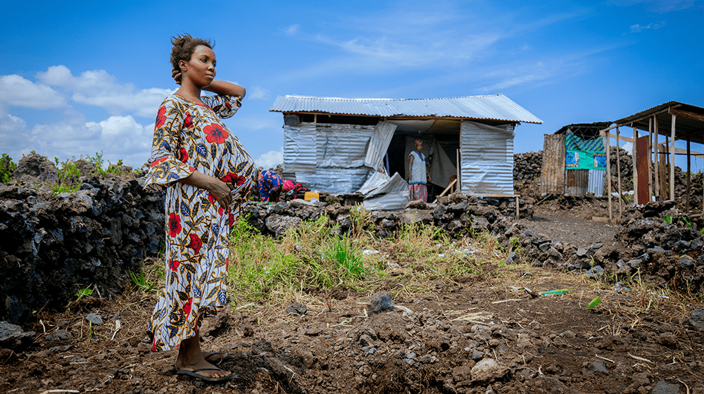 A pregnant woman in a floral dress stands before a shelter made of misshapen corrugated metal.