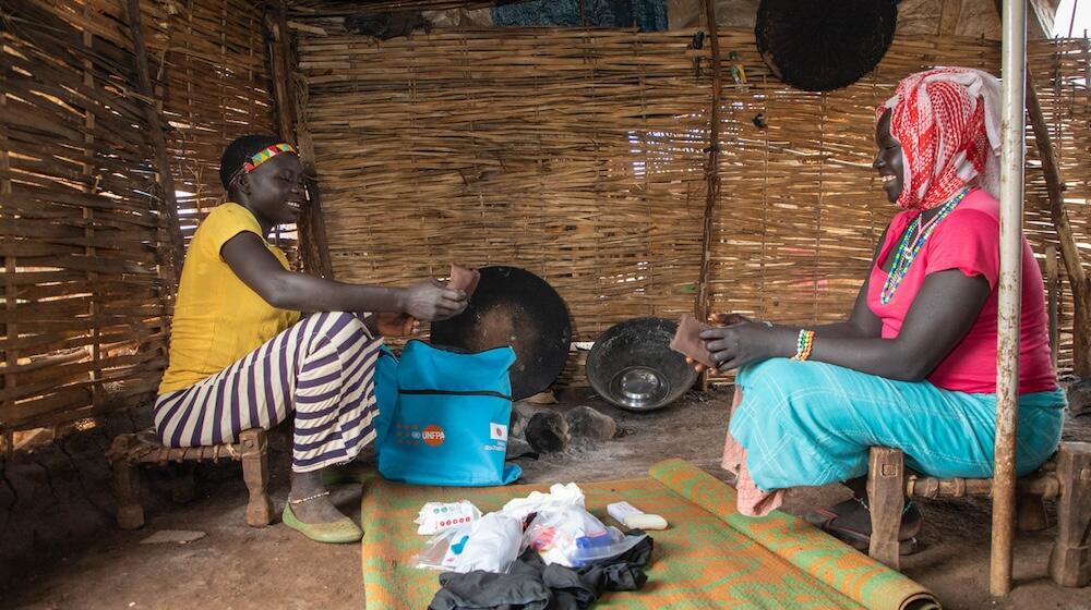 Two girls sit in a hut smiling and looking through dignity kit contents on the floor.