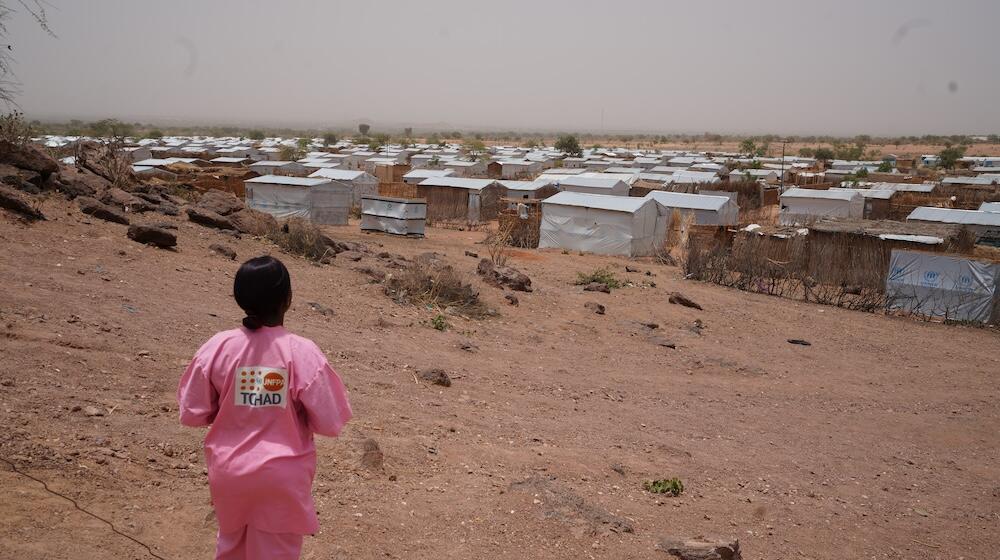 A midwife in a pink uniform walks through an arid landscape dotted with temporary shelters.
